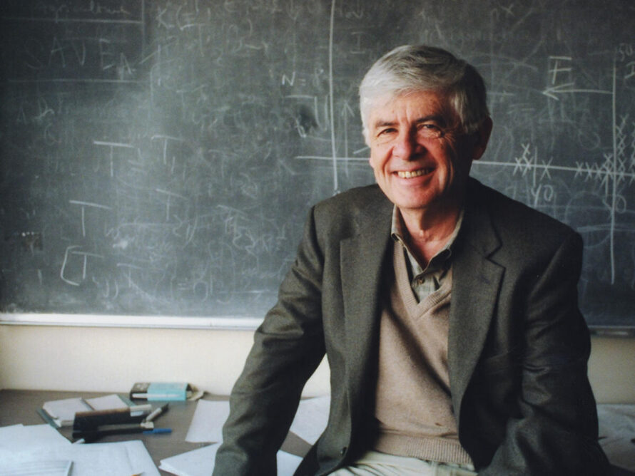 Person wearing jacket and vest sitting in front of blackboard.